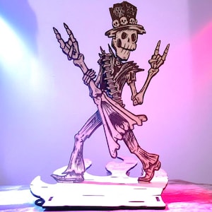 Puede incluir: Un recorte de madera de un esqueleto tocando la guitarra, con un sombrero de copa con detalles de calaveras. El esqueleto está en una pose de rock and roll, con los brazos levantados en un gesto con la mano. El recorte está sobre una base de madera.
