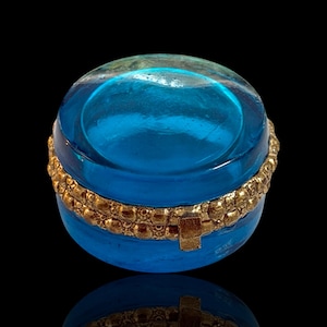 May include: A round, blue glass trinket box with a gold-colored band around the middle. The box has a clear, domed lid. The gold band has a repeating pattern. The box is sitting on a reflective surface.