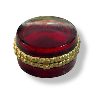 May include: A small, round, red trinket box with a gold-colored band around the middle. The lid is a darker shade of red and has a gold clasp. The box is likely made of glass or a similar material.