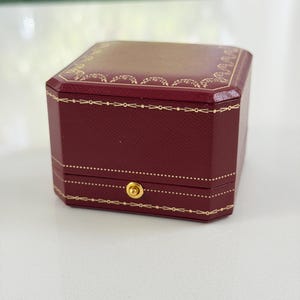 Puede incluir: Una caja de joyería octogonal burdeos con ribetes dorados y un cierre dorado. La caja tiene una superficie texturizada y un interior dorado. La tapa también es burdeos con detalles decorativos dorados.