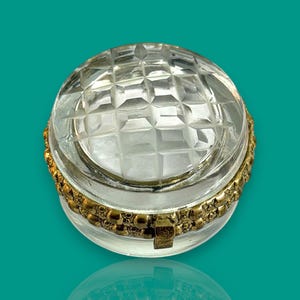 Puede incluir: Una caja de baratijas redonda y transparente con un patrón facetado en forma de diamante en la tapa. La caja tiene una banda de metal dorada alrededor del centro. La caja está sobre una superficie reflectante.