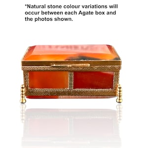 May include: A rectangular box made of orange and red agate stone with a gold-toned metal frame and four small decorative feet. The text above reads: *Natural stone colour variations will occur between each Agate box and the photos shown.