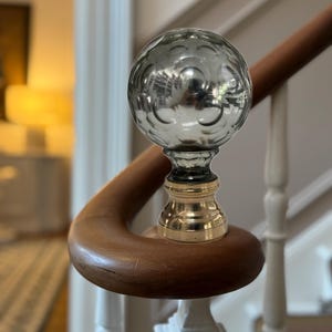 May include: A decorative stair post finial featuring a silver glass orb design. The orb sits on a brass and black base, mounted on a wooden handrail. The handrail curves, with white painted spindles in the background.