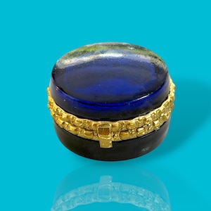 Op de afbeelding: Een kleine, ronde sieradendoos met een donkerblauwe, ovaalvormige deksel. De doos heeft een goudkleurige band rond het midden en een zwarte basis. De deksel heeft een reflecterend oppervlak met een vleugje groen en bruin. Op een helderblauwe achtergrond.