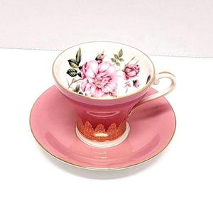 May include: A pink teacup and saucer set with a floral design. The cup has a gold trim and the saucer has a pink rim.