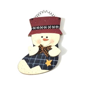 May include: A wooden snowman ornament in the shape of a stocking. The snowman is wearing a red hat with a white snowflake trim, a black and white bow tie, and a blue and white plaid stocking. The stocking has a yellow star on it.