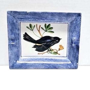 May include: A blue framed ceramic tile with a black bird perched on a branch with green leaves and yellow flowers. The bird has a yellow stripe on its wings.