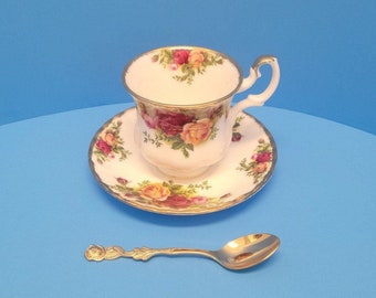May include: A white teacup and saucer with a floral pattern of red, yellow, and pink roses. The cup and saucer have a gold trim. A gold teaspoon is also included.