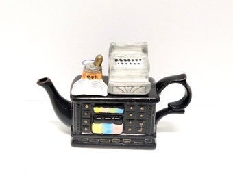 May include: Black teapot shaped like a vintage cash register with a teacup and a book on top. The teapot has gold accents and a black handle.