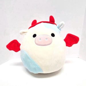 May include: A soft toy cow in cream, with black eyes and a pink nose. It features red horns and wings, and a light blue patch. The toy is designed to be cuddly and is suitable for children.