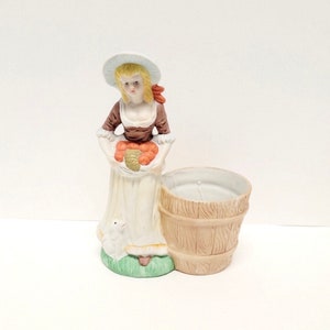 May include: A ceramic figurine of a woman in a white dress and hat, holding a basket of fruit. The figurine is next to a light brown barrel-shaped container. The woman has blonde hair and a red bow. The base has a small lamb on green grass.