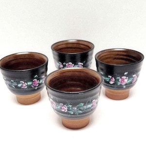 May include: Set of four black ceramic teacups with a brown glaze and hand-painted pink floral designs.