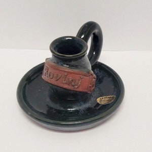 May include: A green ceramic candle holder with a red clay label that says "Rovinj". The holder has a small round base and a handle.