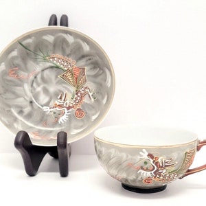 May include: A gray and white teacup and saucer set with a gold rim and a dragon design. The dragon is painted in shades of red, orange, and green.
