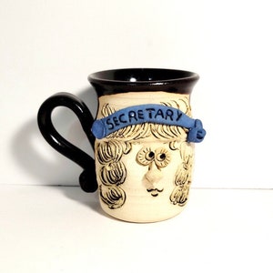 Peut inclure: Une tasse en céramique avec un glaçage brun et un visage blanc avec une bannière bleue qui dit "SECRETARY". La tasse a une poignée noire et un glaçage blanc sur le visage. Le visage a des traits exagérés, y compris de grands yeux, une bouche large et une coiffure bouclée.