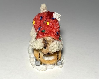 May include: A small, white, fluffy dog wearing a red and green knitted hat is sitting on a wooden sled. The dog is looking down at the sled.