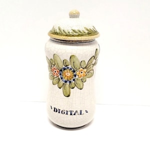 May include: White ceramic jar with a lid, decorated with green leaves and colorful flowers. The jar has a crackled glaze and the word "DIGITAL" written in blue on the front.