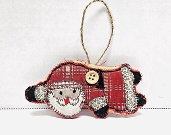 May include: A handmade Christmas ornament featuring a red and white plaid Santa Claus with a white beard and a black button nose. The ornament is made of fabric and has a brown button for a belly button. The ornament is hanging from a brown twine string.