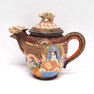 May include: An antique Japanese teapot with a dragon spout and lid finial. The teapot is primarily maroon with detailed artwork featuring figures, gold accents, and intricate patterns. The handle and spout are gold-colored.