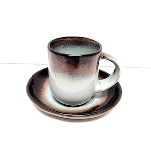 May include: A ceramic coffee cup and saucer set. The cup and saucer have a gradient color scheme, transitioning from dark brown to white. The cup has a rounded shape with a handle. The saucer is round and complements the cup's design.