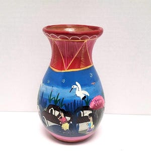 May include: A small, hand-painted ceramic vase with a blue and white scene of a village with people and a bird. The vase has a red rim and a pink base.