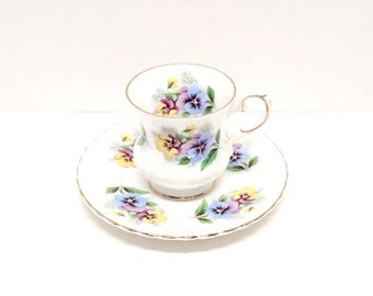 May include: A white teacup and saucer set with a floral pattern of purple, yellow, and blue pansies. The set has a gold trim around the edges.