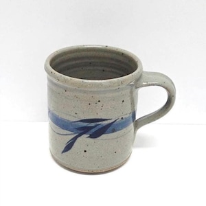 May include: A handmade ceramic mug with a speckled gray finish. A blue band with leaf-like designs wraps around the mug. The mug has a curved handle and is suitable for hot or cold beverages.