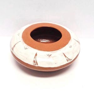 May include: A small, round ceramic bowl with a wide opening. The bowl is primarily white with a brown rim and base. It features brown, stylized, feather-like designs around the exterior. The interior of the bowl is a darker brown.