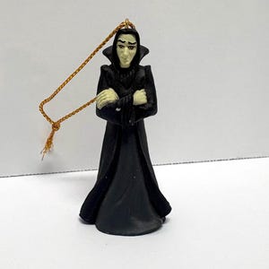 May include: A figurine ornament depicting a character with pale skin, dark hair, and a black hooded robe with a high collar. The figure has arms crossed over the chest and is attached to a thin gold-colored hanging cord for display purposes.