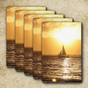 May include: A set of five rectangular wall art prints featuring a golden sunset over the ocean with a sailboat silhouetted against the bright light. The scene shows calm water reflecting the sun, distant mountains, and a clear sky with clouds.