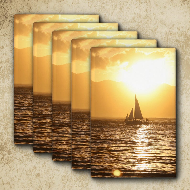 May include: A set of five rectangular wall art prints featuring a golden sunset over the ocean with a sailboat silhouetted against the bright light. The scene shows calm water reflecting the sun, distant mountains, and a clear sky with clouds.