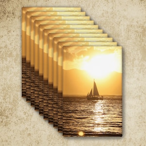 May include: A stack of rectangular wall art prints featuring a golden sunset over the ocean with a silhouette of a sailboat. The scene displays a bright sun low on the horizon, casting a shimmering reflection across the rippling water surface.