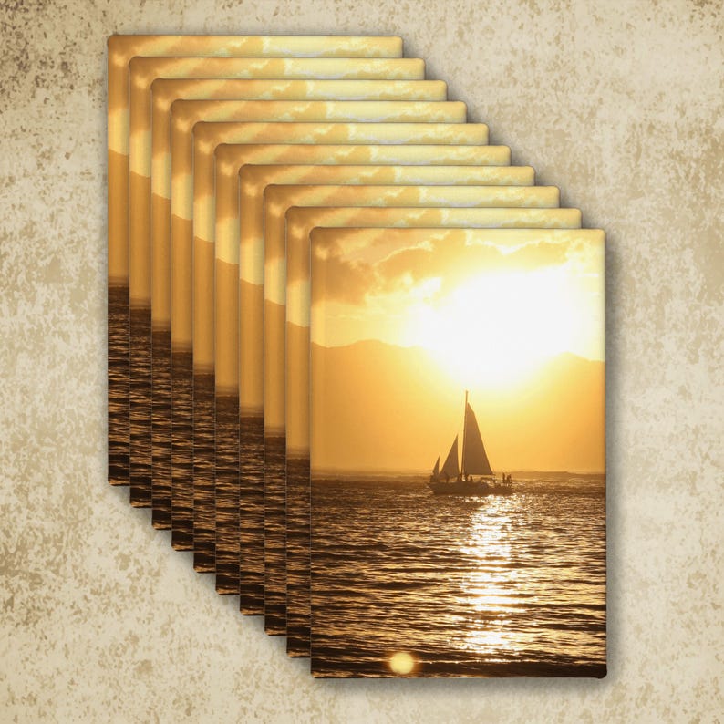 May include: A stack of rectangular wall art prints featuring a golden sunset over the ocean with a silhouette of a sailboat. The scene displays a bright sun low on the horizon, casting a shimmering reflection across the rippling water surface.