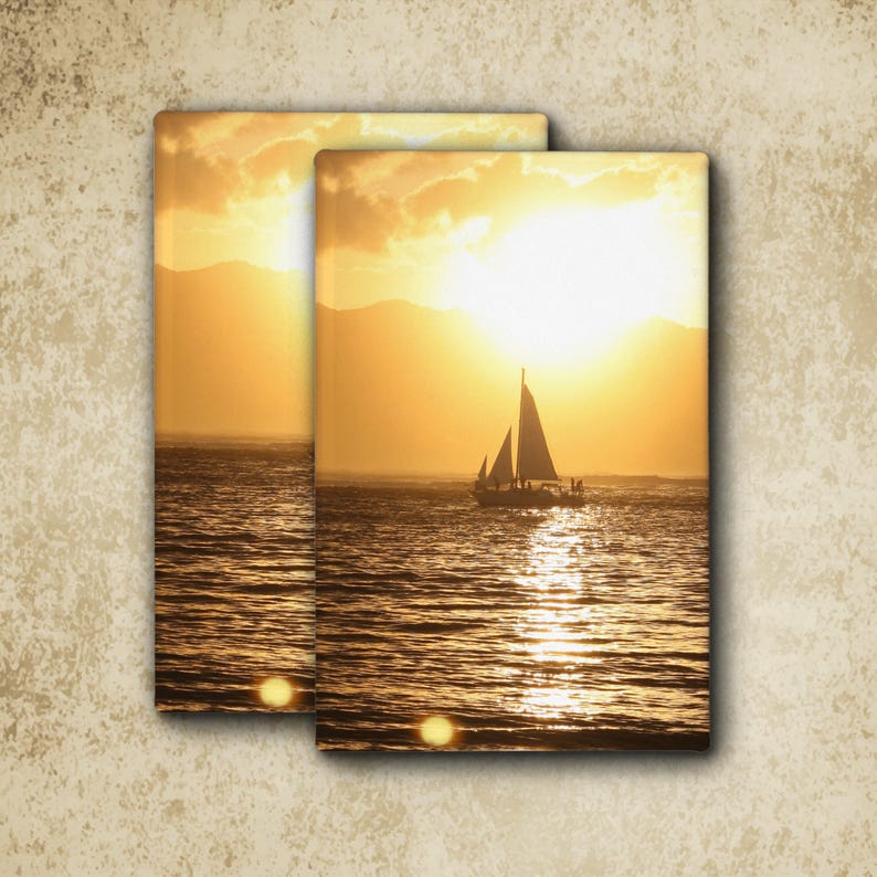 May include: Two rectangular canvas prints featuring a sunset over the ocean with a sailboat silhouette. The scene displays golden light reflecting on the water, distant mountain outlines, and a bright sun positioned behind the sailboat's sails.
