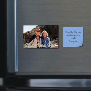May include: A rectangular photo magnet featuring a man in a cowboy hat and a woman standing outdoors in front of rocky terrain. The text "I Love You, Dad" is printed in white script at the bottom. Next to it is a light blue square sticky note.