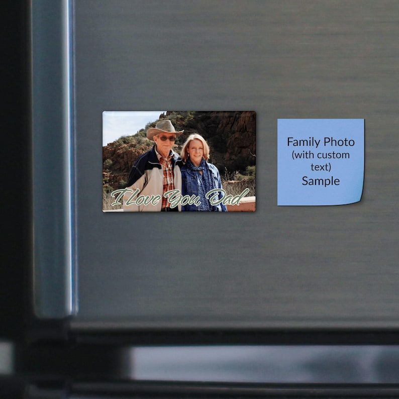 May include: A rectangular photo magnet featuring a man in a cowboy hat and a woman standing outdoors in front of rocky terrain. The text "I Love You, Dad" is printed in white script at the bottom. Next to it is a light blue square sticky note.