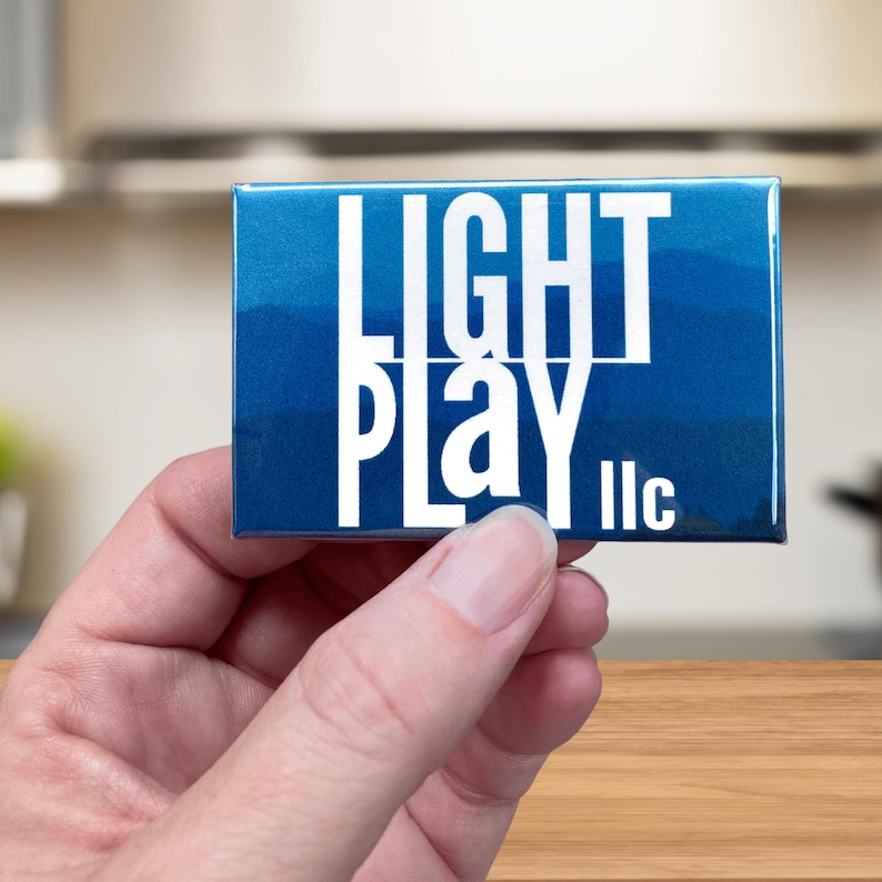 Close up image of Light Play llc branded magnet held by finger tips in front of kitchen counter background.