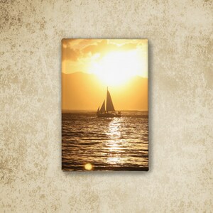 May include: A rectangular canvas print featuring a sailboat silhouetted against a bright golden sunset over the ocean. The sun hangs low in the sky, casting a shimmering reflection across the water's surface, with distant mountains visible in the background.