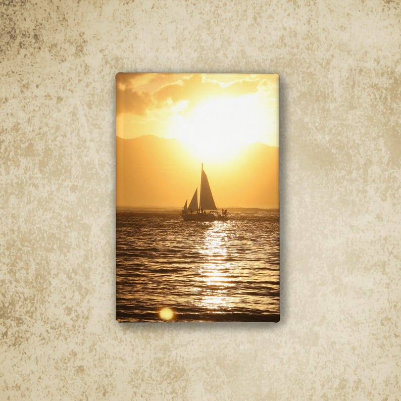 May include: A rectangular canvas print featuring a sailboat silhouetted against a bright golden sunset over the ocean. The sun hangs low in the sky, casting a shimmering reflection across the water's surface, with distant mountains visible in the background.