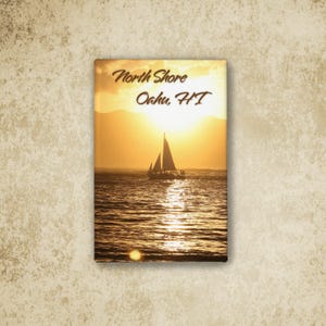 May include: A rectangular magnet featuring a sunset scene over the ocean with a sailboat silhouette. The text North Shore Oahu, HI is printed in dark brown script. The golden sun reflects on the water surface, with mountains visible in the distance.