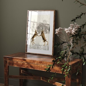 May include: A framed print of a vintage illustration of two brown hares boxing in a field. The background is a muted grey with a tree and a distant landscape.