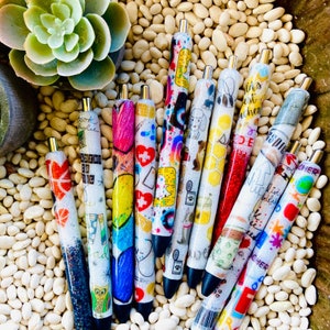 Pu&ograve; includere: Una collezione di 11 penne glitter colorate con vari disegni e testi. Le penne sono disposte a ventaglio su uno sfondo bianco. Il testo sulle penne include "Bookaholic", "Reading is the best teacher", "Life is a little bit of sparkle" e "Teacher".