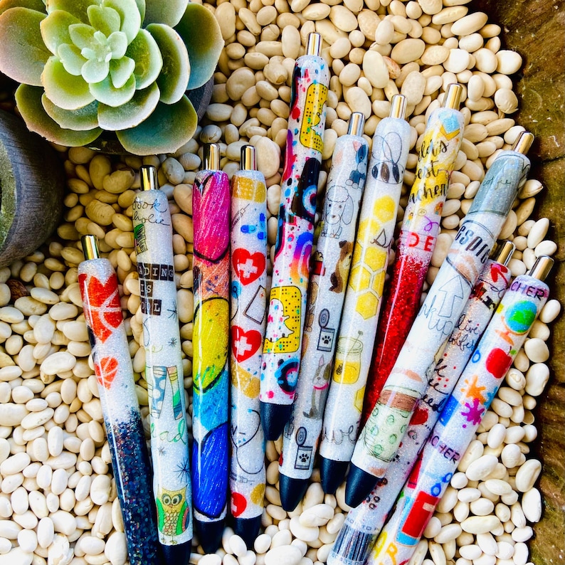 Pu&ograve; includere: Una collezione di 11 penne glitter colorate con vari disegni e testi. Le penne sono disposte a ventaglio su uno sfondo bianco. Il testo sulle penne include "Bookaholic", "Reading is the best teacher", "Life is a little bit of sparkle" e "Teacher".