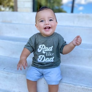 May include: A young child wearing a green short-sleeved shirt with the text "Rad like Dad" in white. The child is wearing blue shorts and gray sneakers. The child is sitting on a set of concrete steps.