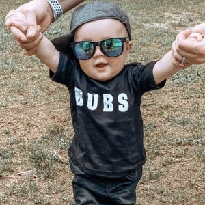 May include: A toddler wearing a black t-shirt with the word "BUBS" printed in white, black jeans, a gray baseball cap, and blue sunglasses. The toddler is wearing black and white sneakers.