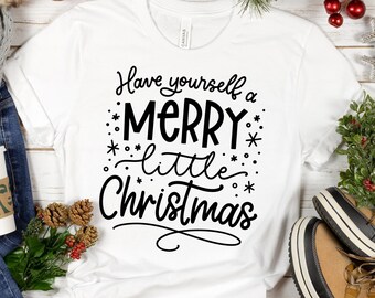 May include: White t-shirt with black text that reads "Have yourself a merry little Christmas". The shirt is on a white wooden surface with pine branches, red berries, and brown boots.