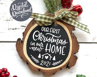 May include: A round wooden ornament with a black chalkboard surface. The ornament features the text "Our First Christmas in our new HOME 2021" with a small house and heart graphic. The ornament is decorated with a green and white plaid ribbon and red bells.