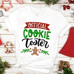 Puede incluir: Camiseta blanca con el texto "OFFICIAL COOKIE TESTER" en letras negras y verdes. Un hombre de jengibre está debajo del texto con hojas verdes y bayas rojas a cada lado.
