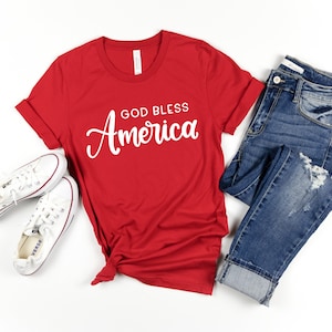 May include: Red t-shirt with the text "God Bless America" in white cursive lettering. The shirt is paired with blue jeans and white sneakers.