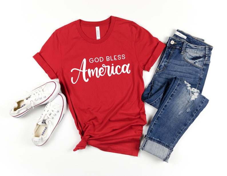 May include: Red t-shirt with the text "God Bless America" in white cursive lettering. The shirt is paired with blue jeans and white sneakers.